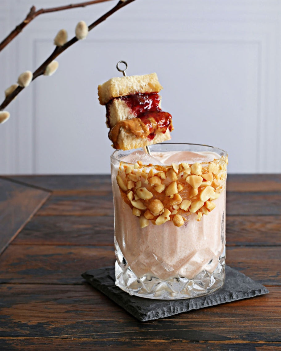 A creamy Peanut Butter and Jelly cocktail with peanut butter whiskey, Rustic Oak Blackberry Mixer, and half and half, garnished with a PB&J sandwich skewer.