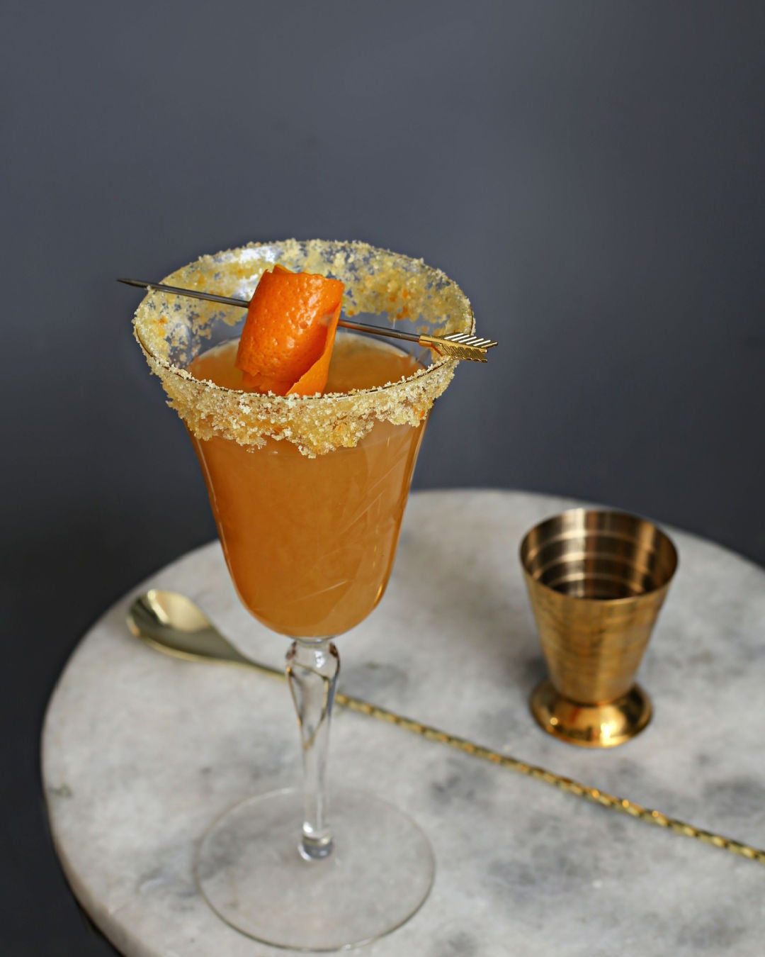 A Sidecar cocktail in a coupe glass with an orange sugared rim and orange peel garnish next to a bottle of Rustic Oak Orange Barrel Aged Mixer on a marble surface.