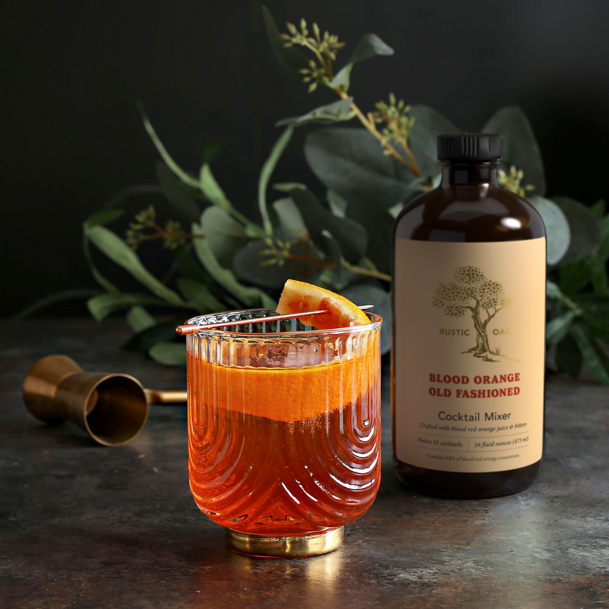 A styled cocktail scene with a glass of orange-red drink garnished with an orange slice and metal strainer, next to a bottle labeled Rustic Oak Blood Orange Old Fashioned Cocktail Mixer, with eucalyptus leaves and a jigger in the background.
