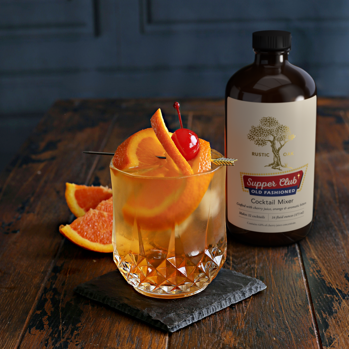 A rustic amber cocktail in a faceted crystal glass with orange slices, a cherry, and a cocktail pick, styled on a dark wooden table next to a bottle of Rustic Oak 'Supper Club' Old Fashioned Cocktail Mixer.